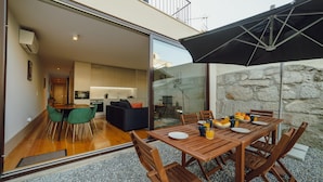 An open-plan living area with a dining table set for breakfast, a kitchen visible through glass doors, and a stone wall outside. - Legacy Oporto Premium Apartment B by Vacationy (Porto)