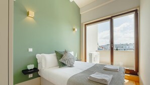 A bedroom with a bed, pillows, a bedside table, a small shelf, and a window with a view of buildings. - Legacy Oporto Premium Apartment I by Vacationy (Porto)