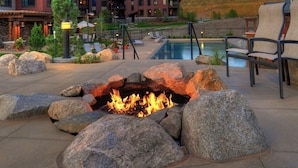 A fire pit surrounded by large rocks, a pool, lounge chairs, and a building in the background. - First Tracks Condo w/ Gondola Access & Pool 1054 (Steamboat Springs)