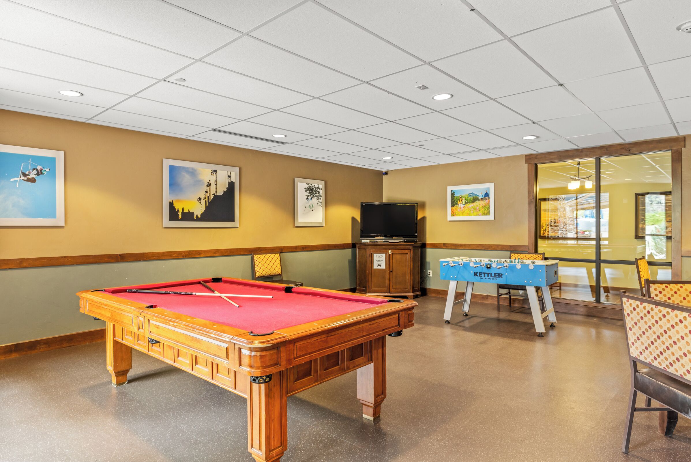 A pool table with a red felt, a foosball table, and a television in a room with beige walls and a tiled ceiling.