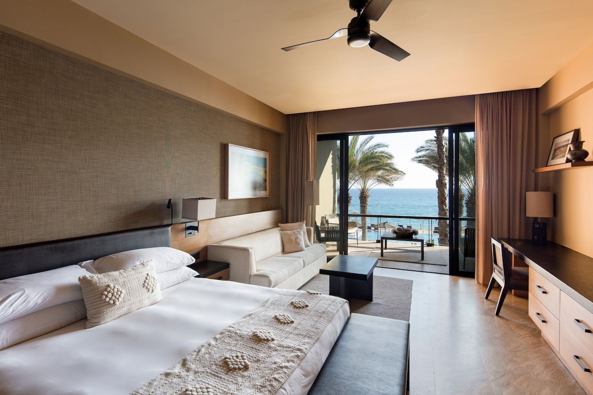 A modern hotel room with a large bed, a sofa, a desk, and a balcony with a view of the sea.