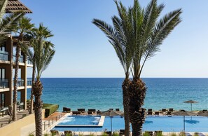 Vista a la piscina con una vista despejada del océano, palmeras y un edificio con balcones.