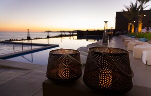 Dos candelabros metálicos con superficie reflectante en una terraza con vistas a una piscina y al mar al atardecer.