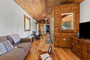 Ein gemütliches Blockhaus-Interieur mit Holzbalkendecke, einer Couch, einem Essbereich und einem Fernseher auf einem Holzschrank.