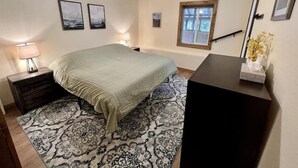 Um quarto com cama, mesa de cabeceira, escrivaninha, ventilador de teto, janela e um tapete estampado.