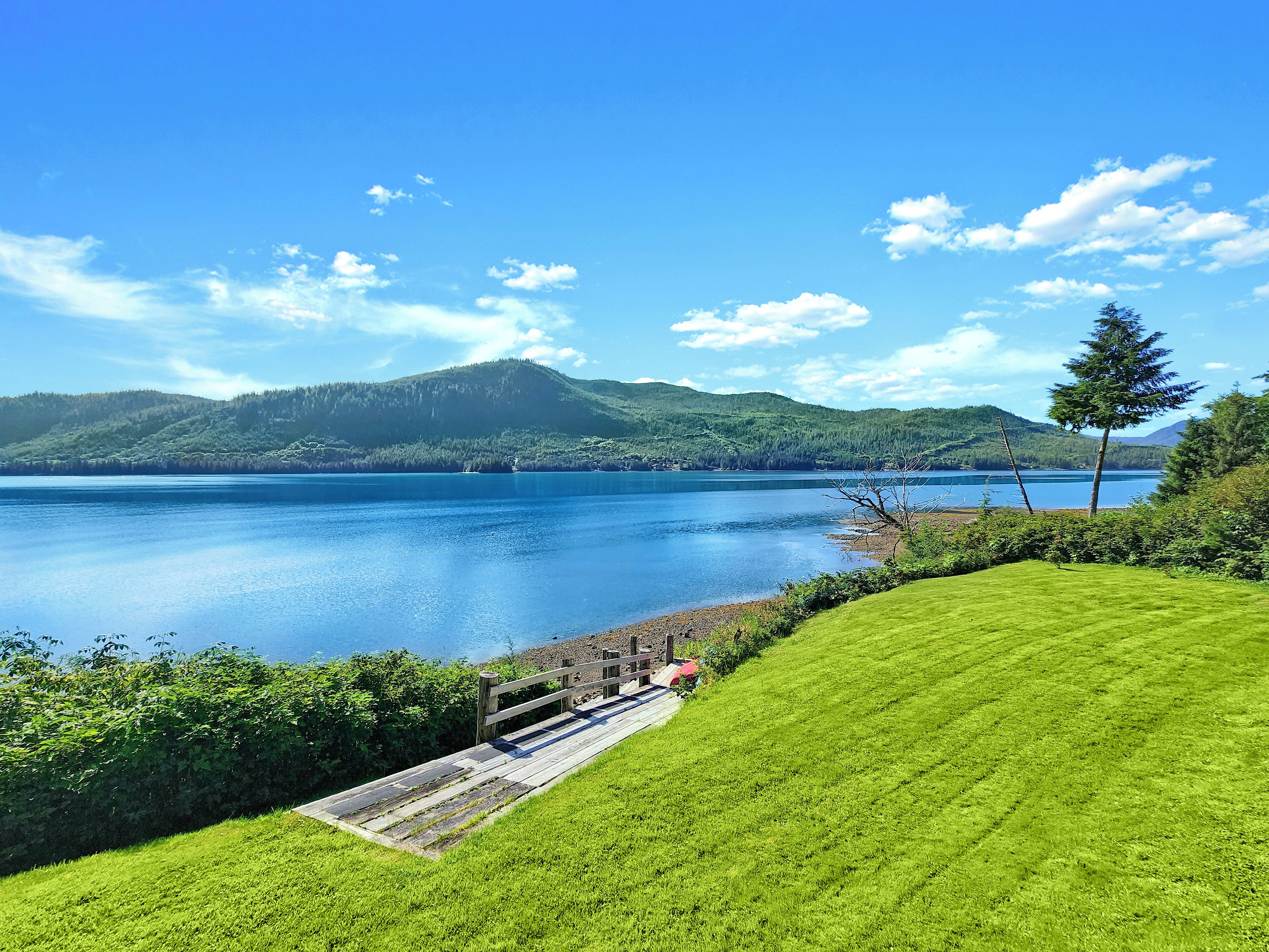 Vista serena de um lago, com gramado, deck de madeira e montanhas ao fundo.