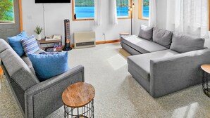 A spacious living room with a large gray sofa, a smaller gray sofa, and a coffee table. There is a flat-screen TV mounted on the wall, a ceiling fan, and large windows offering a view of a lake. - Private 2 bedroom Oceanfront Oasis with Panoramic views! (Craig)