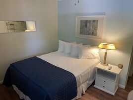A neatly made bed with white linens and a dark blue quilt, a bedside lamp, a small bedside table with a remote control, and a framed picture on the wall.