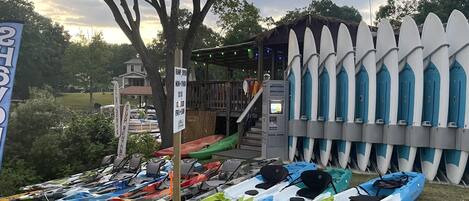 Kayaks alineados junto a un edificio, con un cartel que indica una tarifa de 10 dólares por horarios no convencionales.