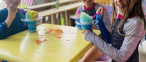 Three children sitting at a yellow table with ice cream cones, under a covered area with a view of trees and an American flag in the background.
