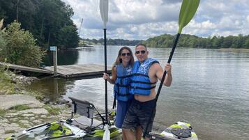 Una pareja de pie junto a un kayak en un muelle, con un lago y árboles al fondo.