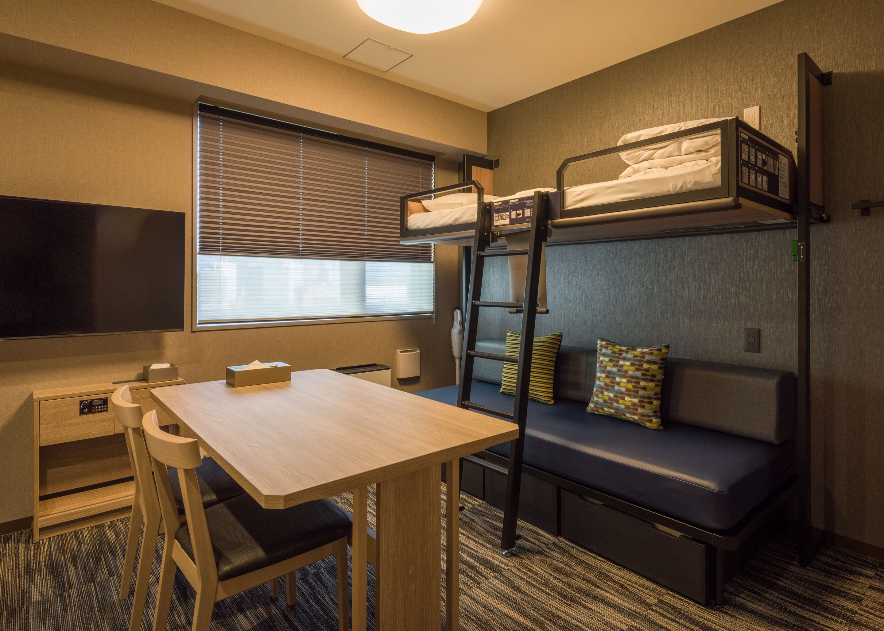 Een kleine hotelkamer met een stapelbed, een bureau met stoel, een televisie en een raam met zonwering.