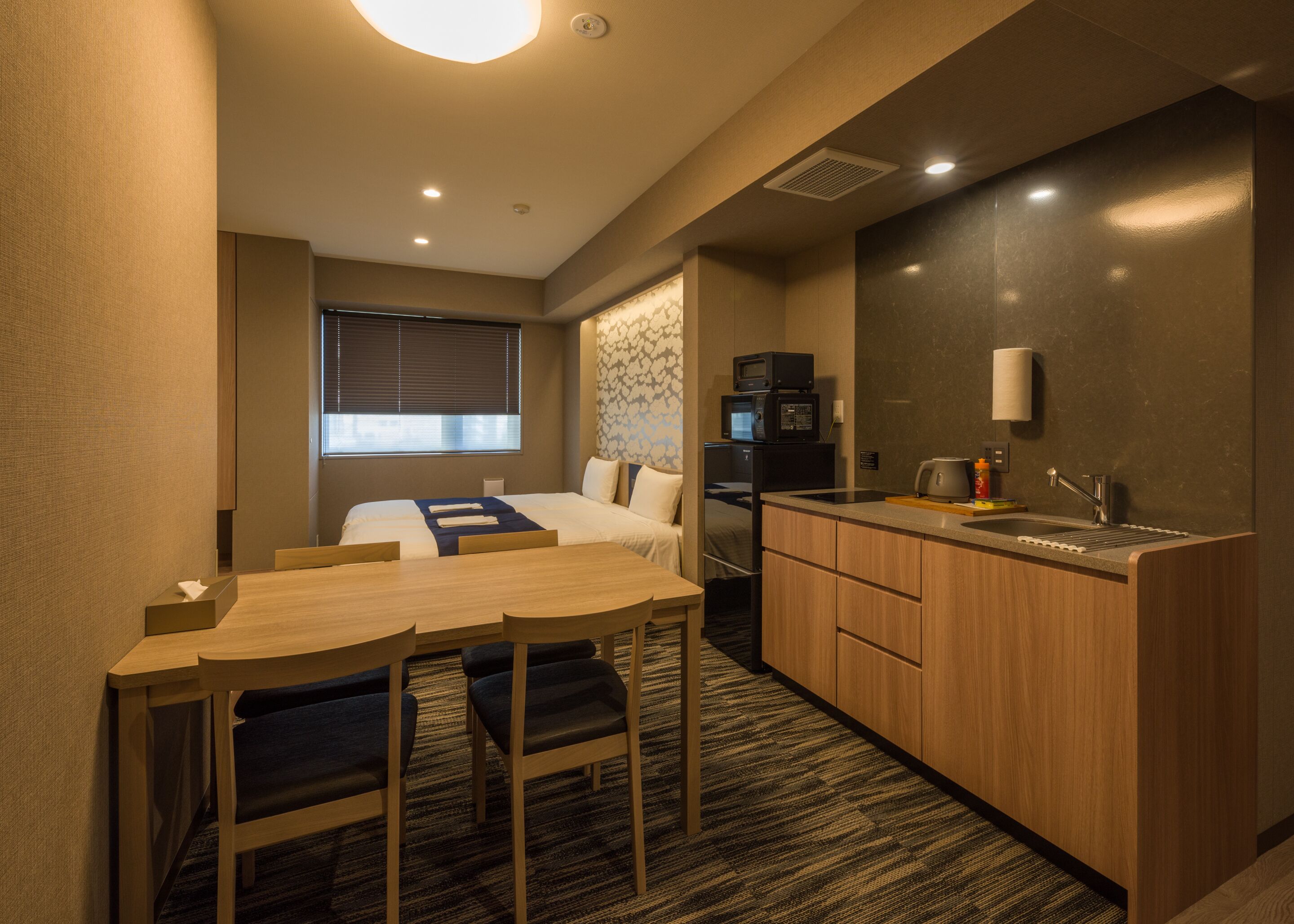 Une chambre d’hôtel compacte comprenant un lit, une petite kitchenette, un coin repas avec une table et des chaises, et une fenêtre avec des stores.