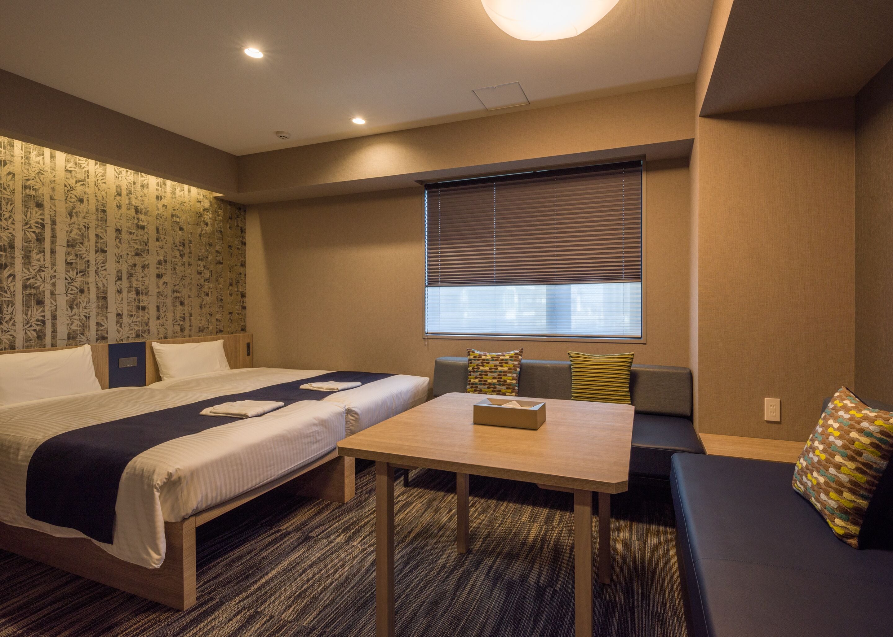 Ein Hotelzimmer mit zwei Betten, einem kleinen Tisch, einer Couch und einem Fenster mit Jalousien.