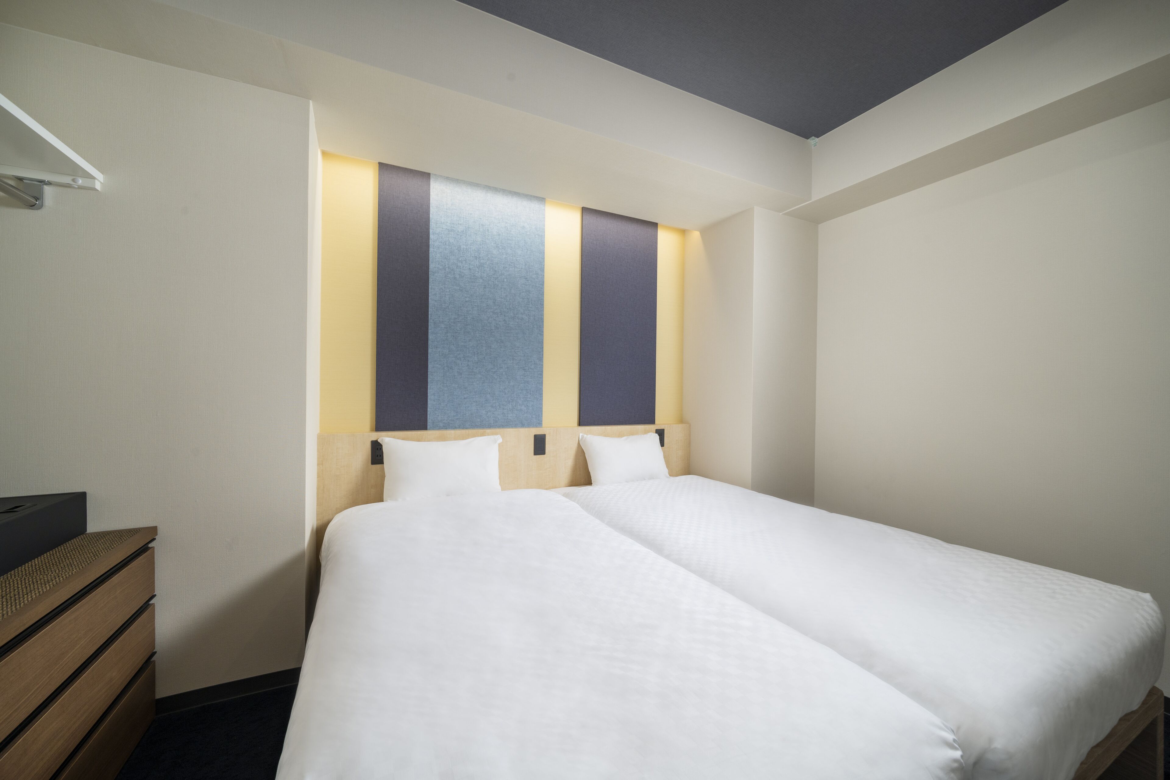 A hotel room with two single beds, a wooden dresser, and a wall-mounted shelf.