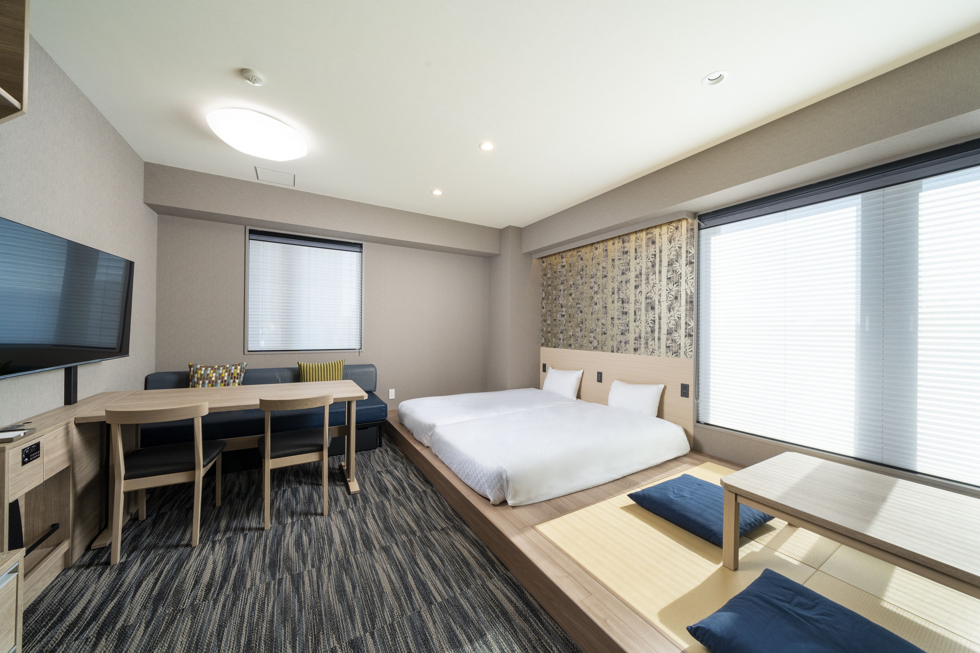 Een moderne hotelkamer met een groot bed, een bureau met stoelen, een flatscreen televisie en een groot raam met jaloezieën.