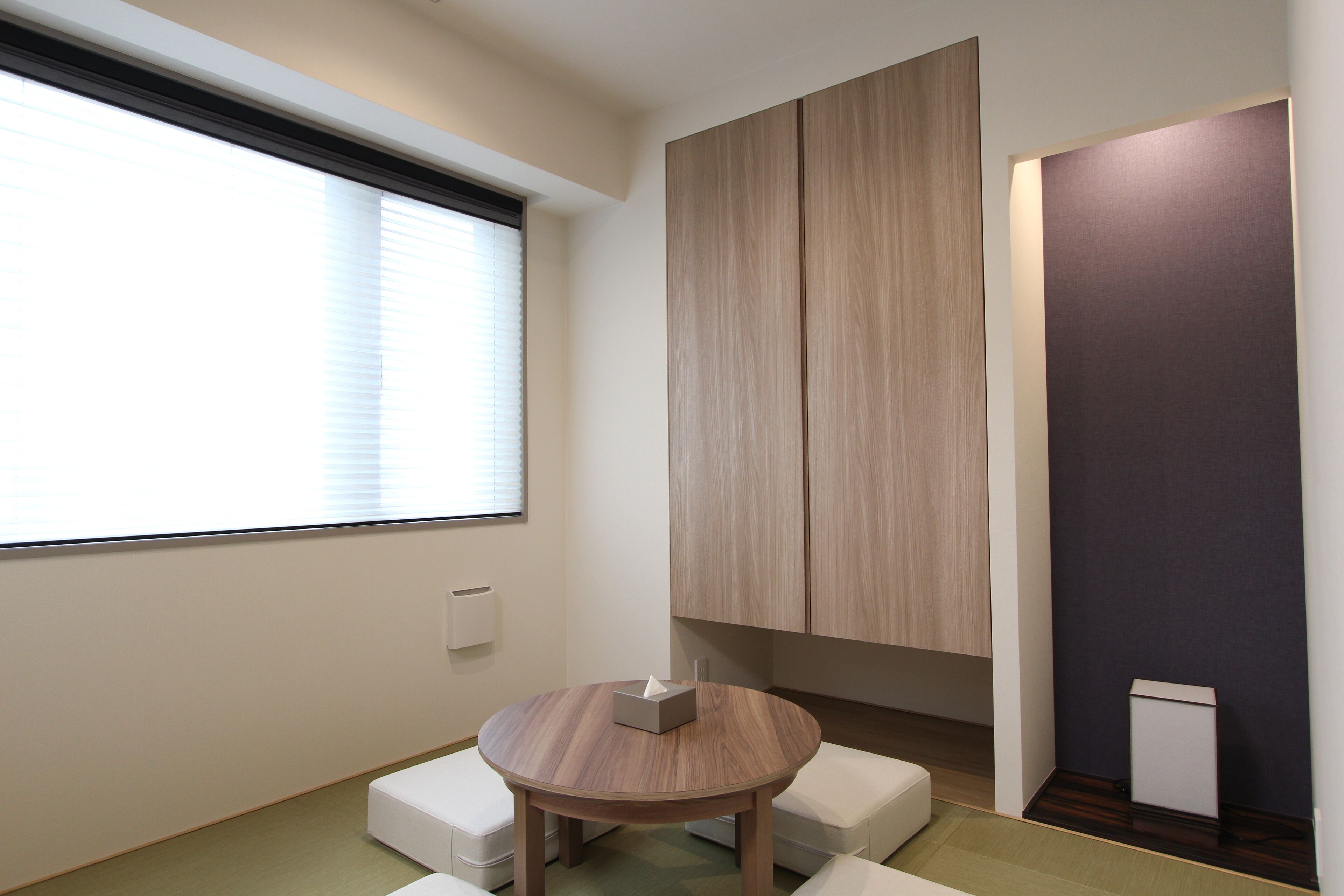 A room with a round table, a window with blinds, a built-in cabinet, and a small white box on the table.