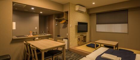 Habitación de hotel con cama, escritorio con silla, kitchenette pequeña, televisión y ventana con persianas.