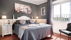 Quarto com cama, mesas de cabeceira, uma cadeira, uma janela grande e um quadro floral na parede.