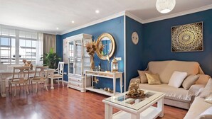 Uma sala de estar com parede azul, sofá bege, mesa de centro branca e um espaço de jantar com cadeiras e armário.