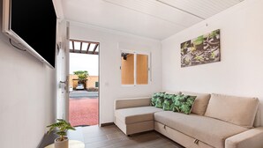 Um quarto com um sofá bege, uma planta em vaso, uma televisão presa à parede e uma porta que dá para o exterior.
