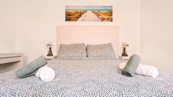 Een slaapkamer met een bed, kussens, een schilderij van een strand en een nachtkastje met een lamp.