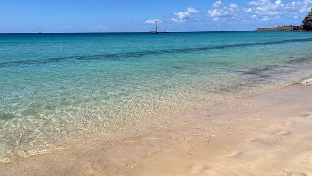 Ein Strand mit klarem türkisblauem Wasser, einem sandigen Ufer und einem entfernten Segelboot.