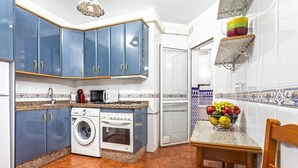 Une cuisine avec des meubles de couleur bleue, un lave-linge, un four, un four à micro-ondes et un panier de fruits posé sur le plan de travail.