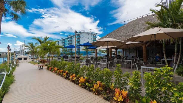 A wooden walkway lined with tropical plants, outdoor seating with umbrellas, and a view of a building with a thatched roof. - 3BR Townhouse (Merritt Island)