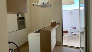 Cozinha moderna com fogão a gás, forno e um refrigerador pequeno. Há um balcão de madeira com uma cesta e um armário branco. Uma porta de vidro deslizante dá acesso a uma varanda com mesa e cadeiras.