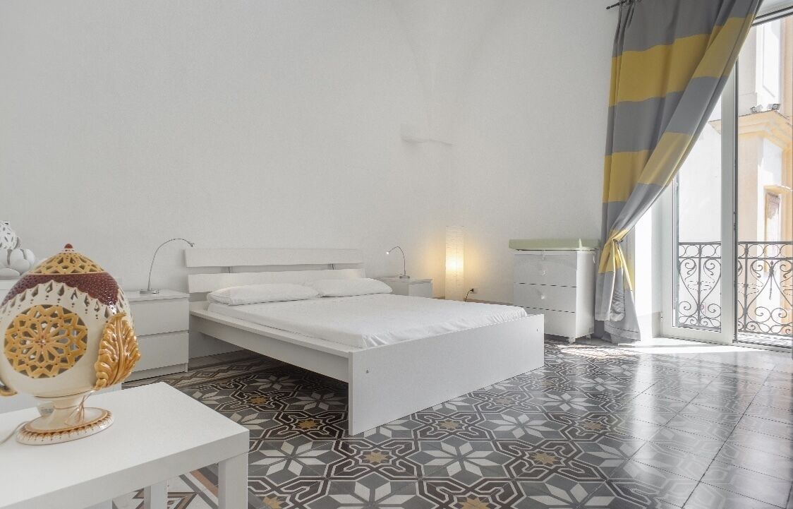 Una camera da letto con un letto bianco, un comodino, una lampada e una ciotola decorativa.