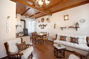 Una sala de estar con sofá, sillas, una mesa de centro, una mesa de comedor y una zona de cocina con televisor y estanterías.
