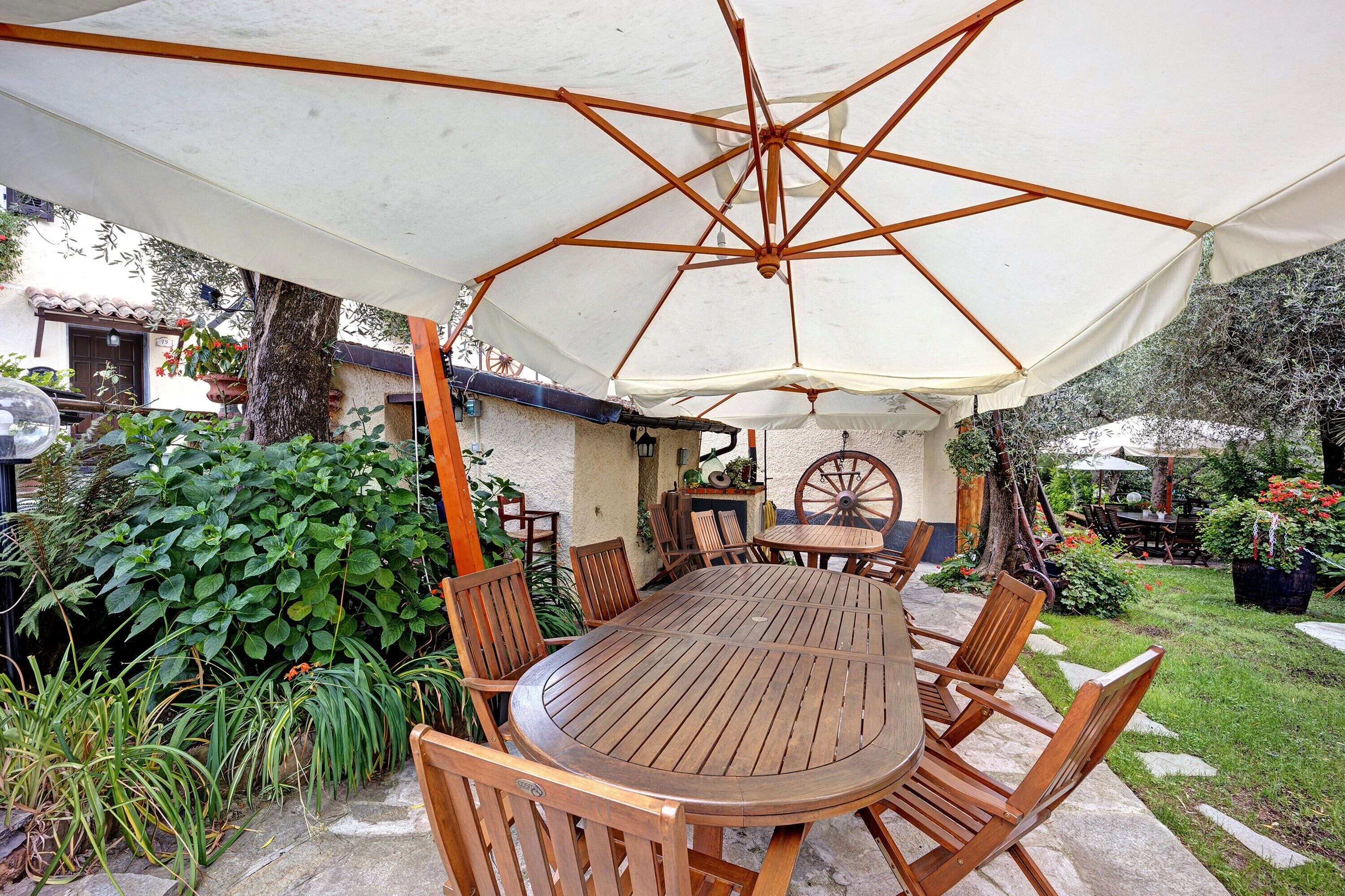 A covered patio with a wooden table and chairs, surrounded by greenery and a small garden.