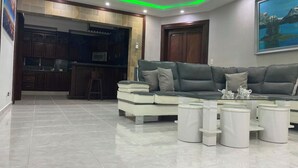 A modern living room with a grey sectional sofa, a white coffee table, and a kitchen area with wooden cabinets and a microwave.