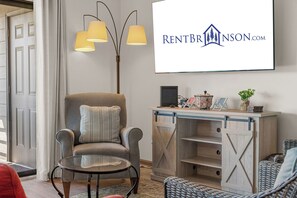 Ein Wohnzimmer mit einem großen Bildschirm, der RENTBRINSON.COM zeigt, einem grauen Sessel mit Kissen, einem kleinen runden Tisch und einer Holzkommode mit Regalen.