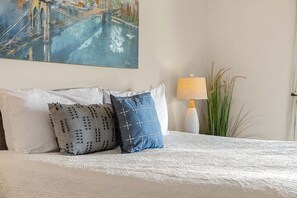Een bed met witte beddengoed, twee decoratieve kussens, een schilderij van een brug, een lamp en een hoge plant.
