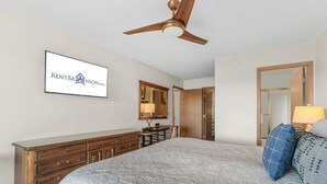 Une chambre à coucher avec un lit, une commode, un miroir, un ventilateur de plafond et un panneau accroché au mur.