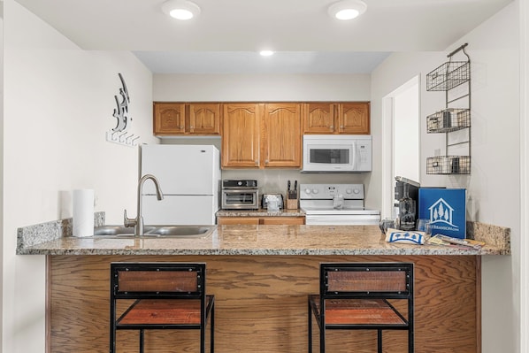A kitchen with wooden cabinets, a stainless steel refrigerator, a microwave, an oven, and a granite countertop. There are two bar stools and a blue sign on the countertop. - Amazing Lake View Walk-In Condo, Newly Renovated Spacious Near Silver Dollar City with Free Tickets (Branson)