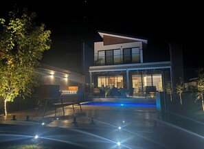 Ein modernes, zweistöckiges Haus mit Swimmingpool, Sitzgelegenheiten im Freien und einer gut beleuchteten Terrasse.
