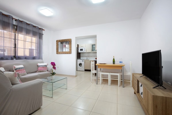 A living room with a sofa, coffee table, TV, and a kitchen area in the background.