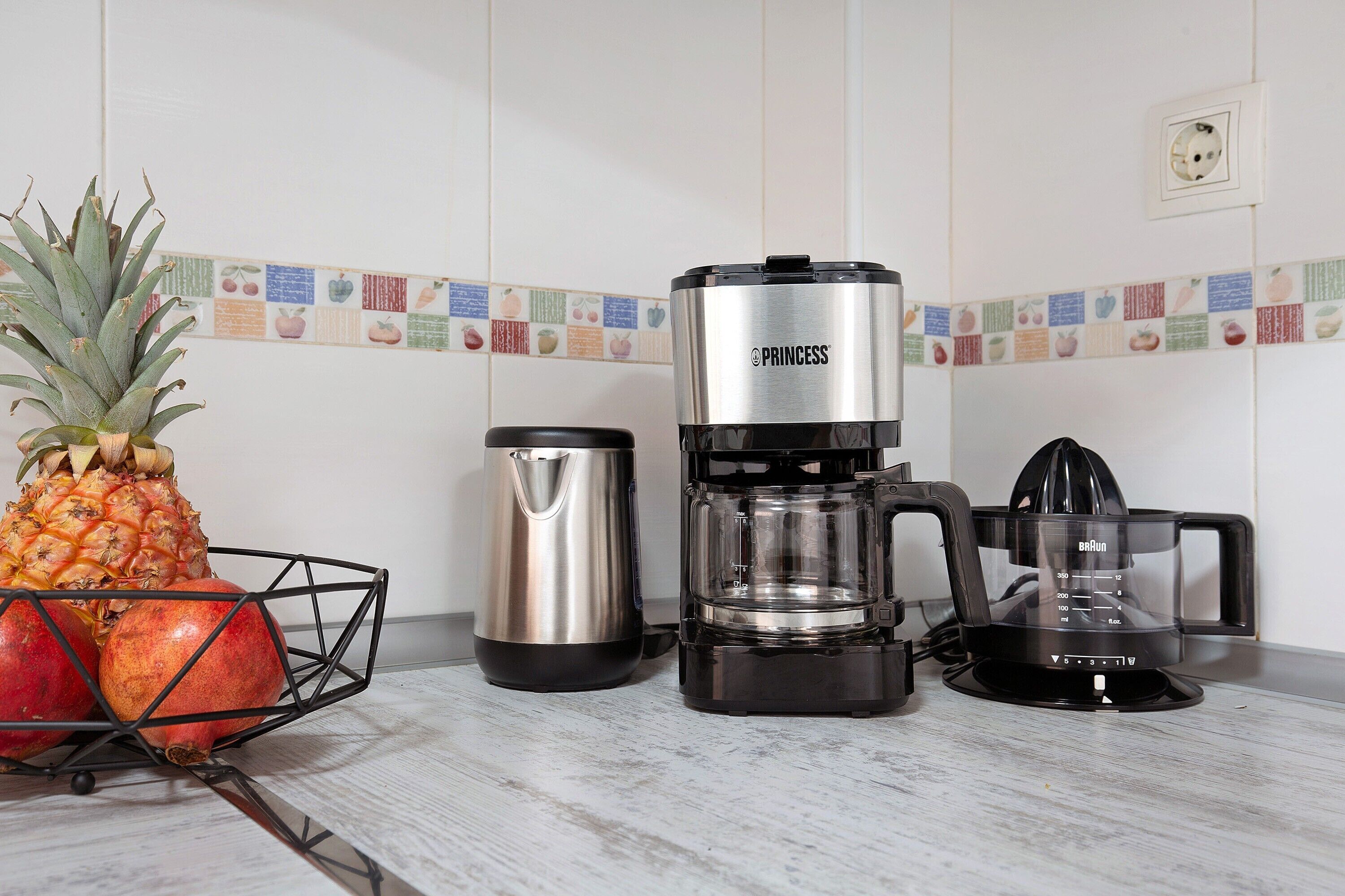 Una cocina con cafetera, hervidor y frutero que contiene una piña y una granada.