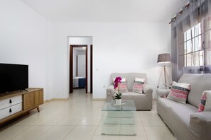 A modern living room with a flat-screen TV, a glass coffee table, a beige sofa, and a lamp.