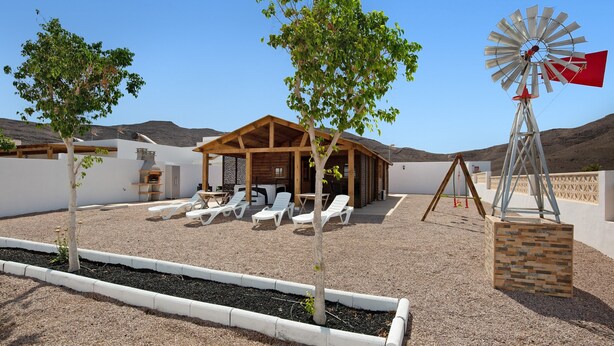A rustic outdoor area with a wooden structure, white lounge chairs, a windmill, and a gravel ground. - Holiday home La Cabañita de Javi with Sea View, Garden & Wi-Fi (Tuineje)