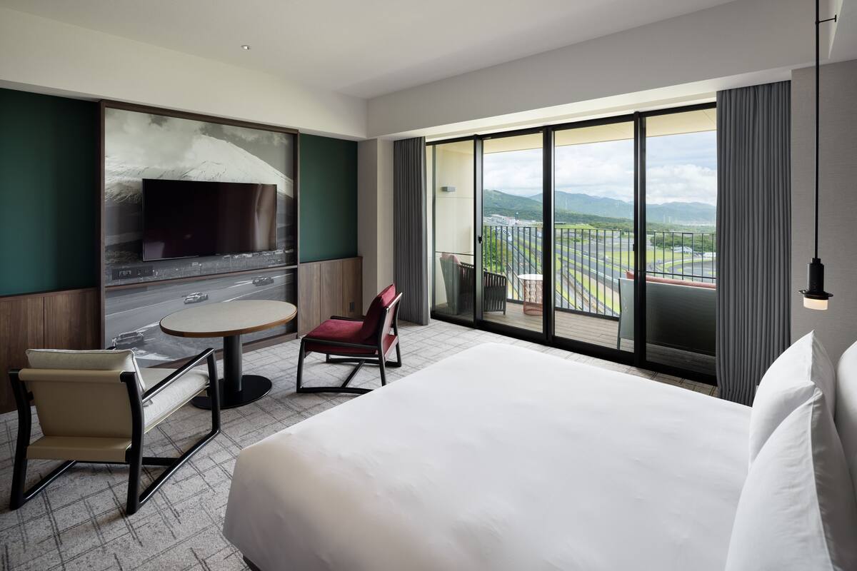 A hotel room with a large bed, a flat-screen TV mounted on the wall, a small round table, a red chair, and a balcony with a view of the city and mountains.