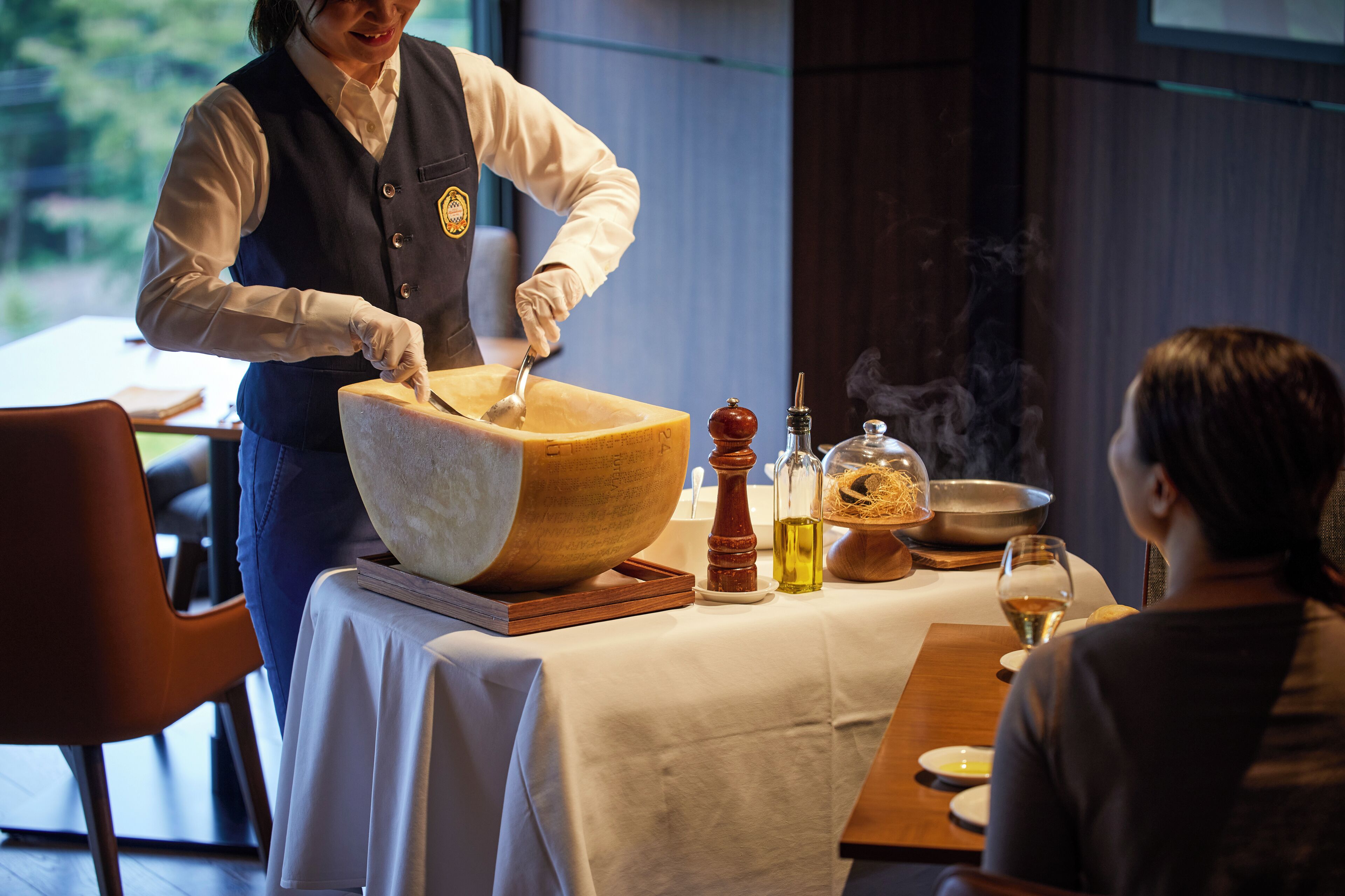 A person preparing food at a table with a large wooden bowl, a pepper grinder, a bottle of oil, and a glass of wine.