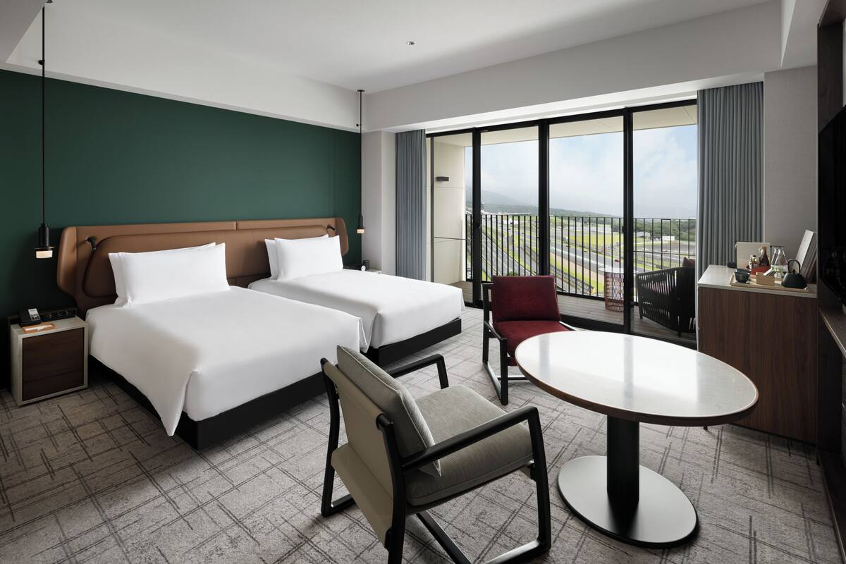 A hotel room with two beds, a desk, a chair, and a balcony with a view.