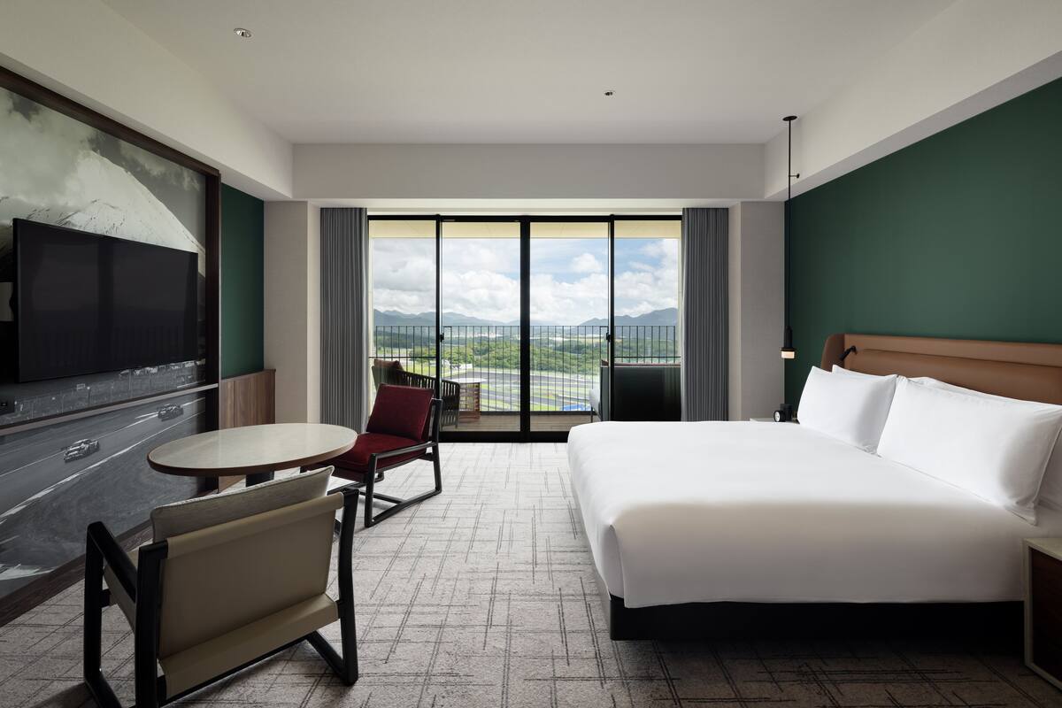 A hotel room with a bed, a TV, a chair, a small table, and a large window with a view of the landscape.