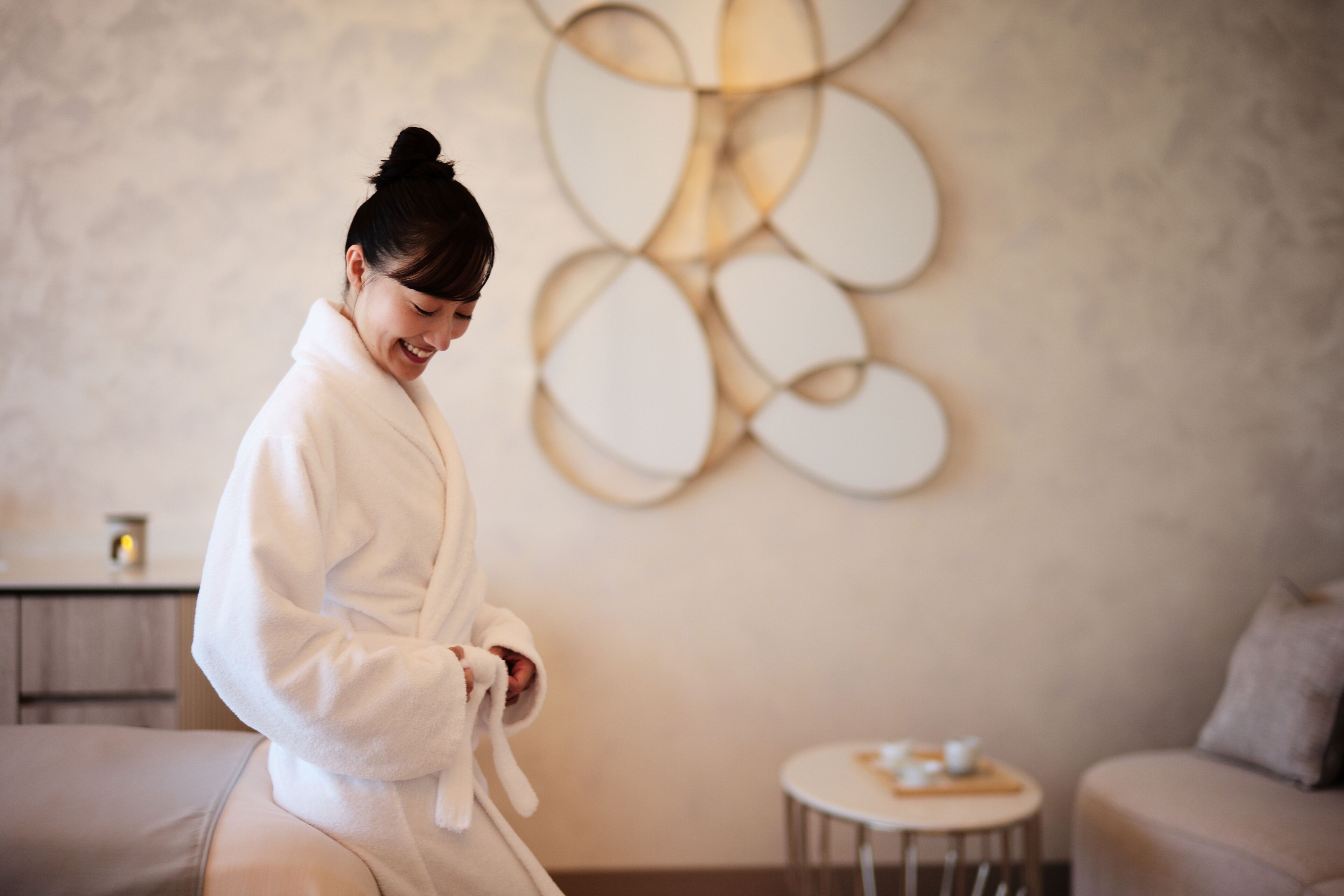 A person in a white bathrobe sitting on a massage table in a room with a modern abstract wall art, a small round table with a cup, and a sofa in the background.