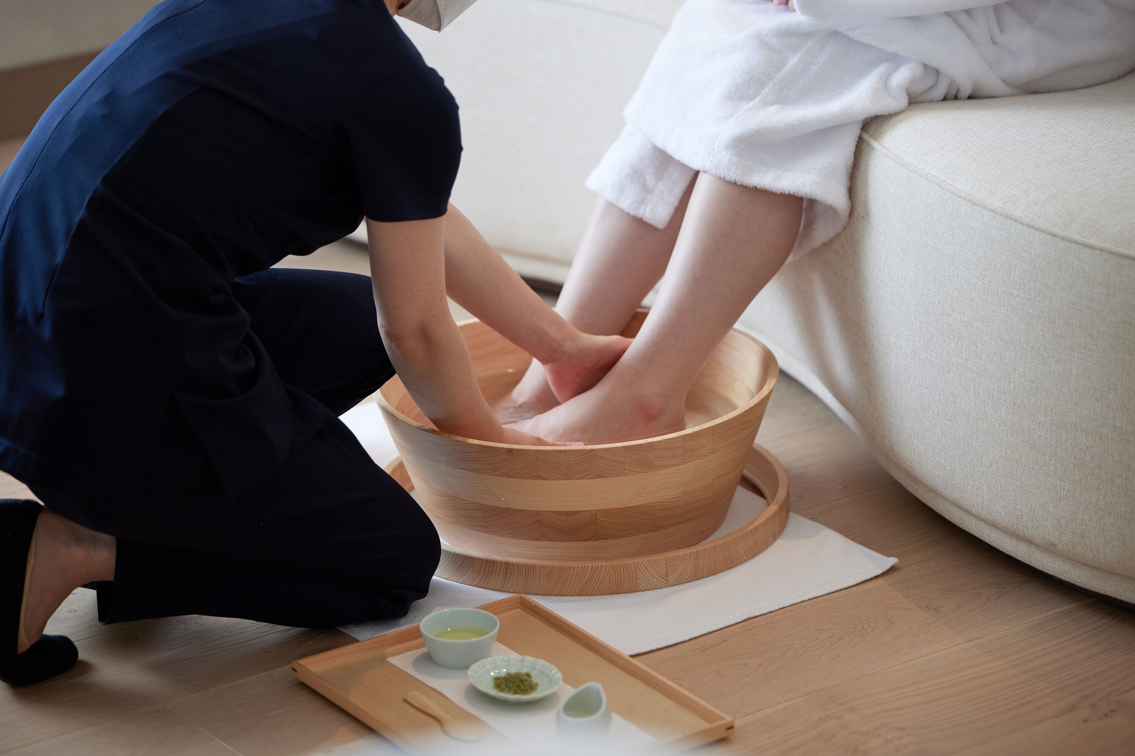 A person receiving a foot soak in a wooden basin on a mat, with a tray of bowls nearby.