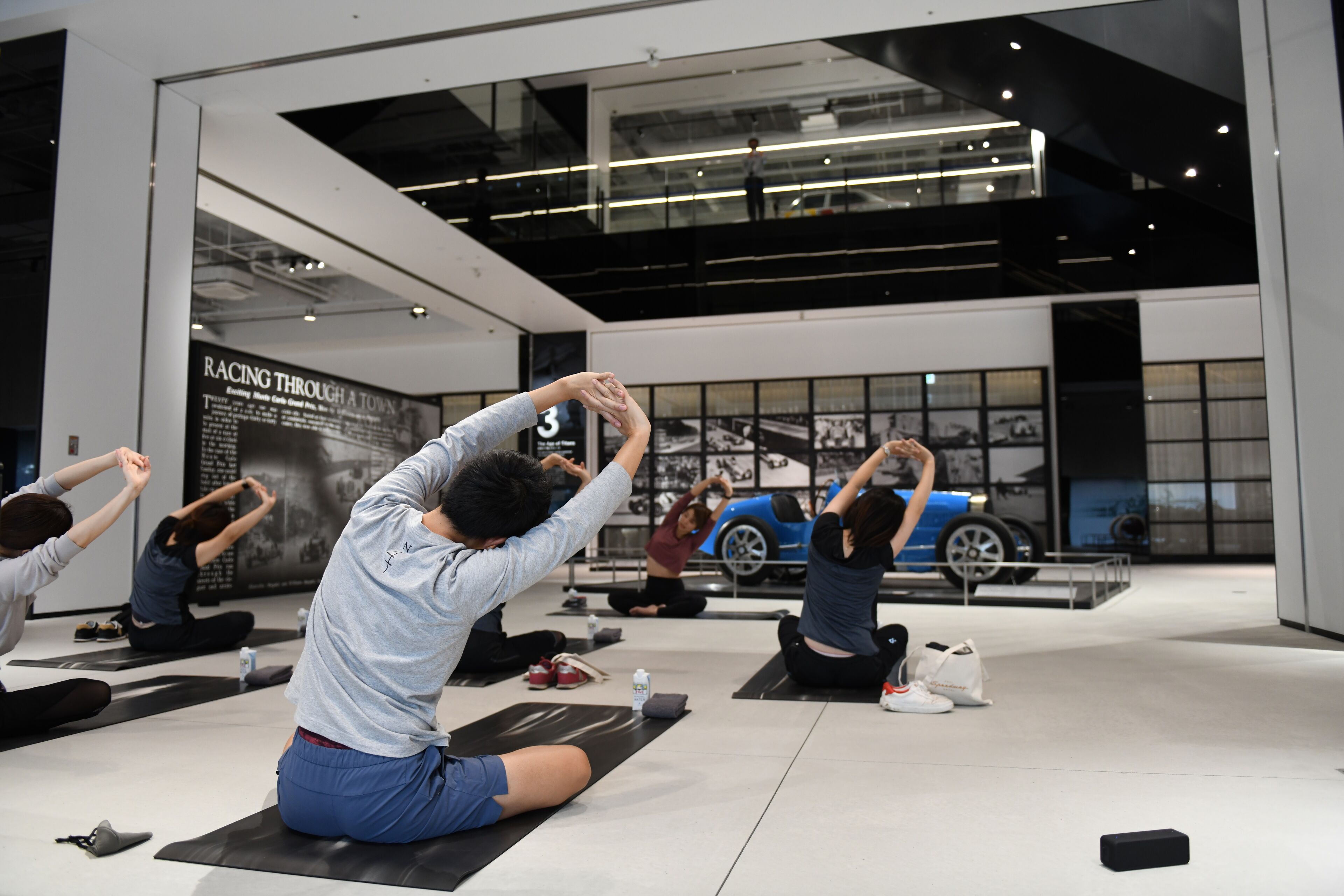 A modern indoor space with people performing yoga on mats, a large digital display, and a blue car model in the background.